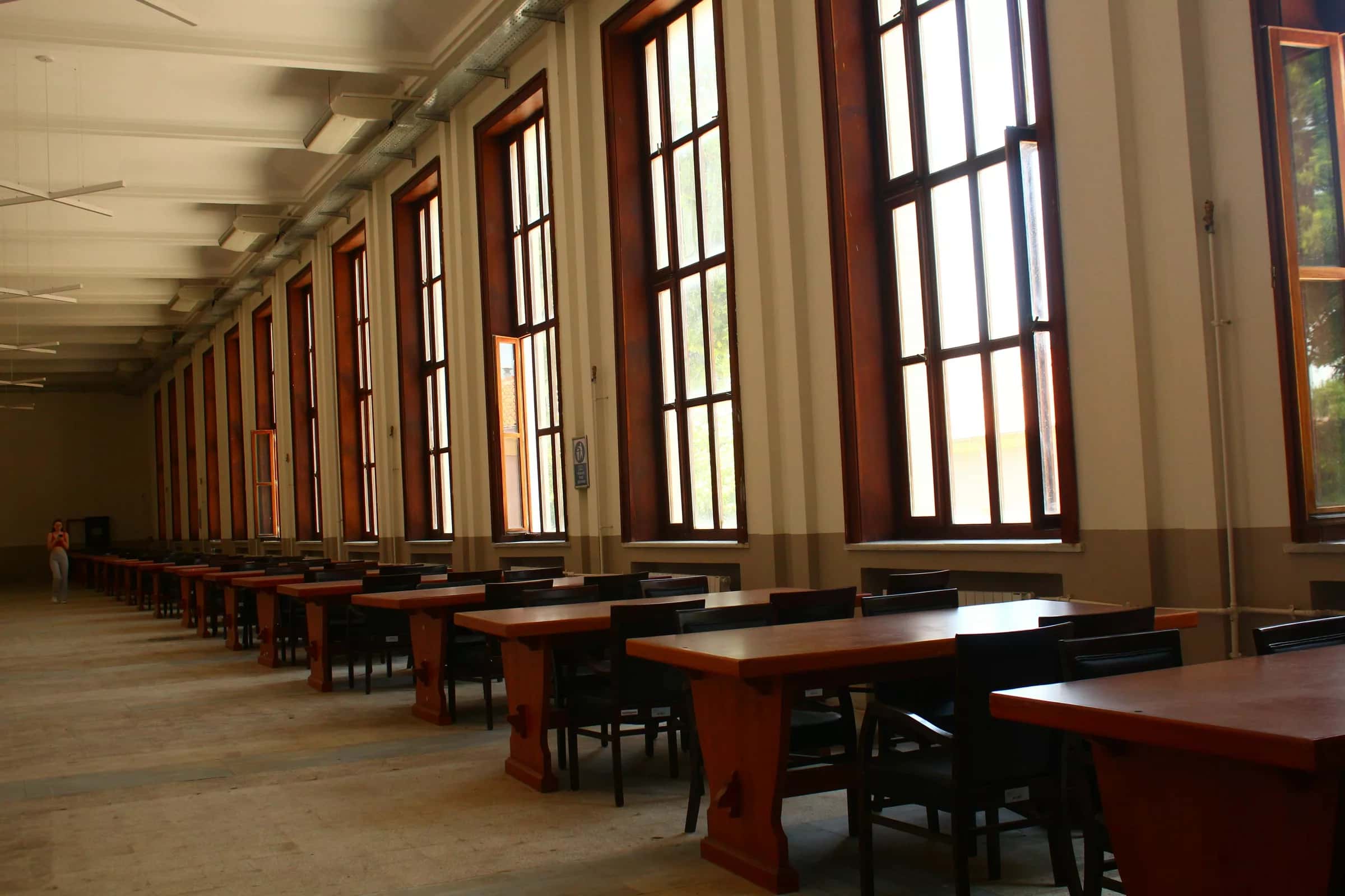 Yüksek pencereli salonda sıra sıra ahşap masa ve sandalyeler